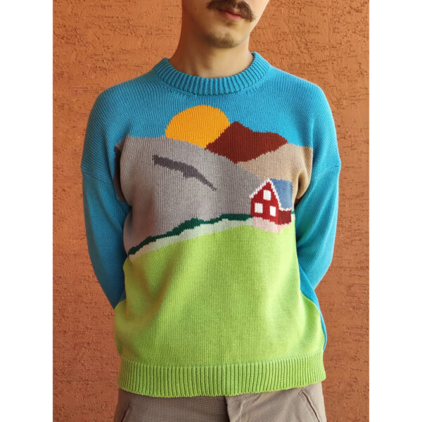A young man standing in front of an orange wall, wearing his ''Nuuk'' landscape sweater which features a house by the mountains and the green fields.