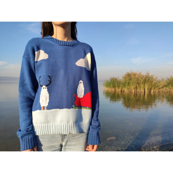 A young woman standing by the lake, wearing her christmas themed intarsia sweater which features two ghosts, one wearing antlers and the other one in a sleigh, alluding to Santa Claus and Rudolph.