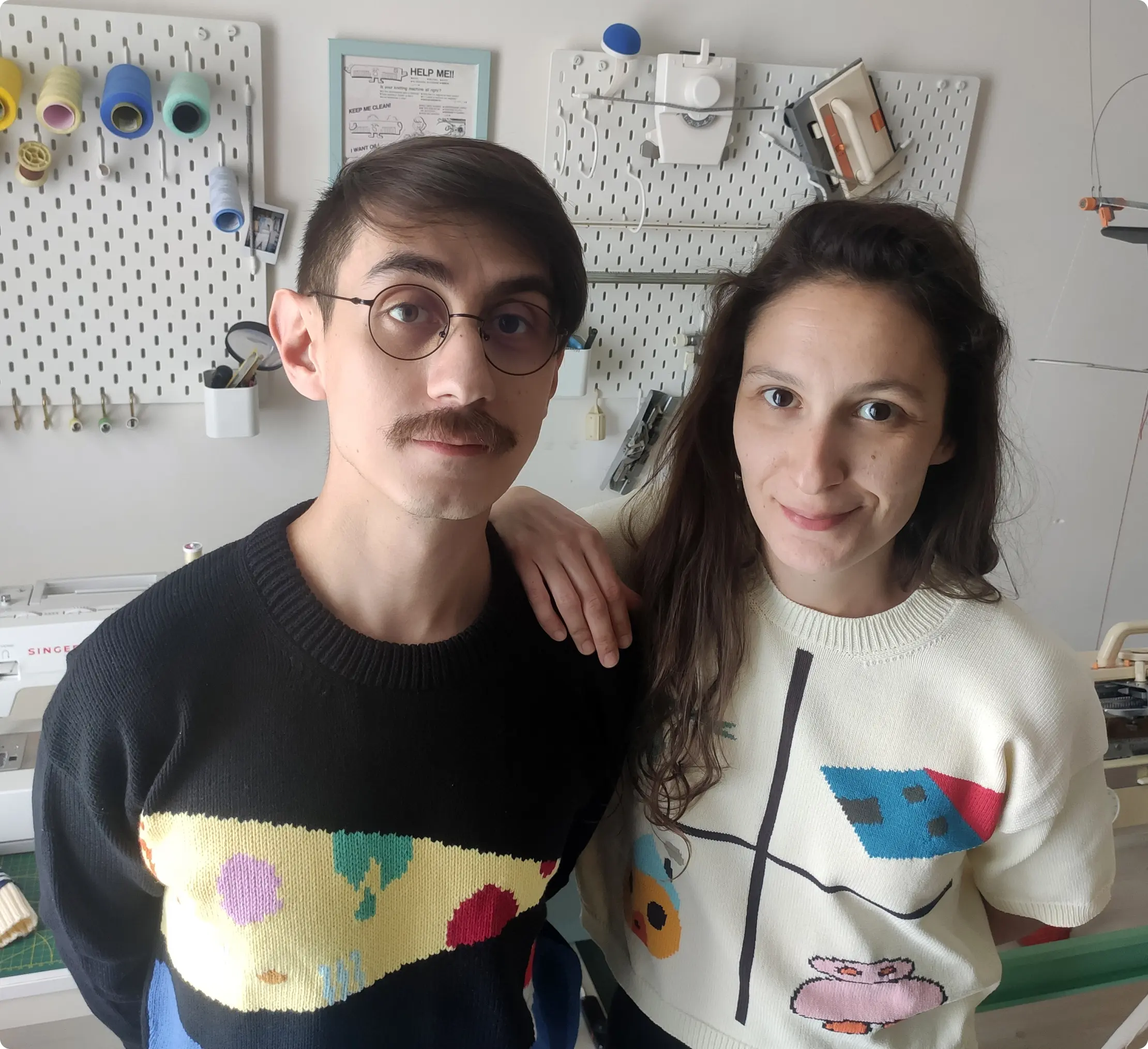 Knitting machine duo Knyt in their studio by the their knitting machine, with various equipment hang on the walls.