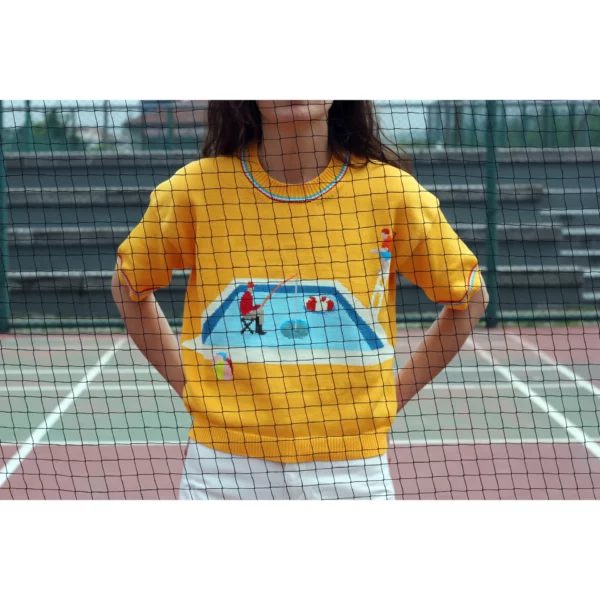 A young woman wearing a mainly yellow coloured knit t-shirt, with a frozen swimming pool, a fisherman and a lifeguard on his post on it. Shot from behing a tennis court net.