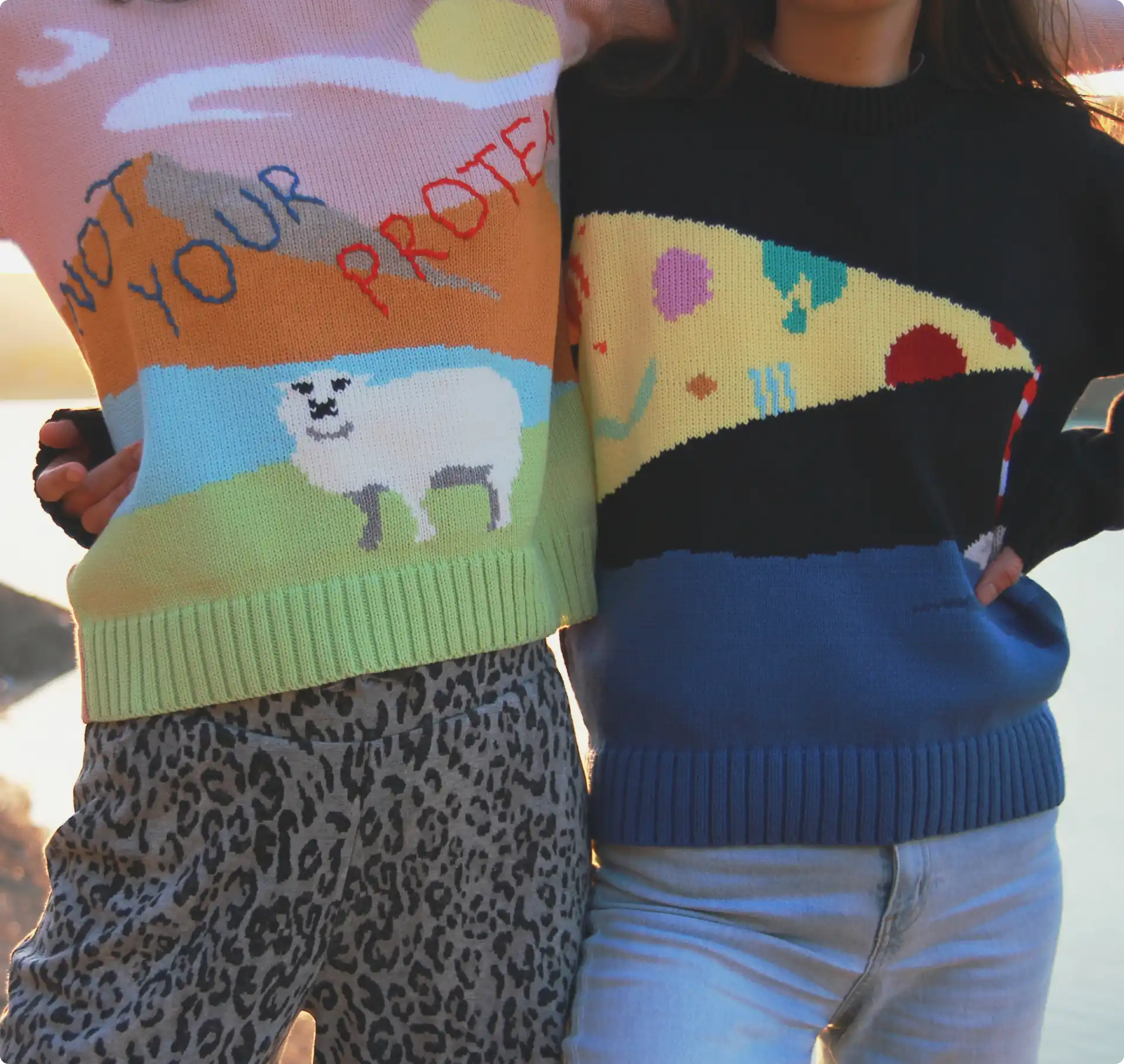 Two models standing by the lake wearing their knit sweaters, one depicting a lighthouse on a dark night and the other a sheep on the hills.