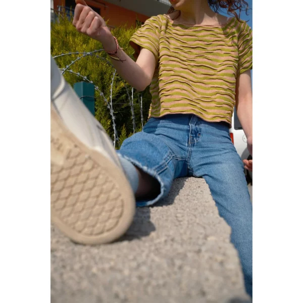 A young woman is seen sitting on a wall wearing Breezer (Caramel), a handmade knit t-shirt with a bumpy texture, crafted with a unique knitting technique in warm tones of Soft Caramel and Chartreuse, made by Knytworks.