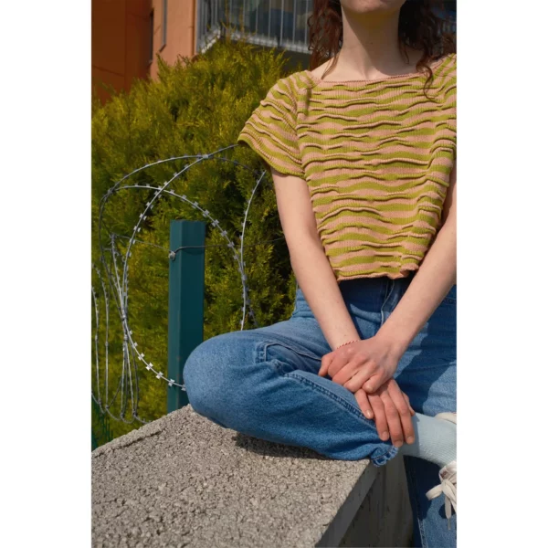 A young woman is seen sitting on a wall, by a tree, wearing Breezer (Caramel), a handmade knit t-shirt with a bumpy texture, crafted with a unique knitting technique in warm tones of Soft Caramel and Chartreuse, made by Knytworks.