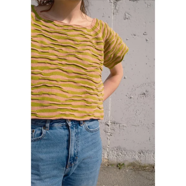 A young woman stands with her arms behind her, wearing Breezer (Caramel). This shot showcases the handmade knit t-shirt, emphasizing the bumpy texture and warm tones of Soft Caramel and Chartreuse.