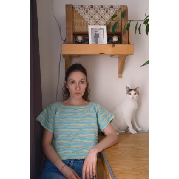 A young woman wearing Breezer (Mint) sits at a table with a cat by her side, both gazing at the camera. The handmade knit t-shirt’s bumpy texture and warm tones of Minty Ice Blue and Muted Olive are beautifully highlighted in this cozy moment.
