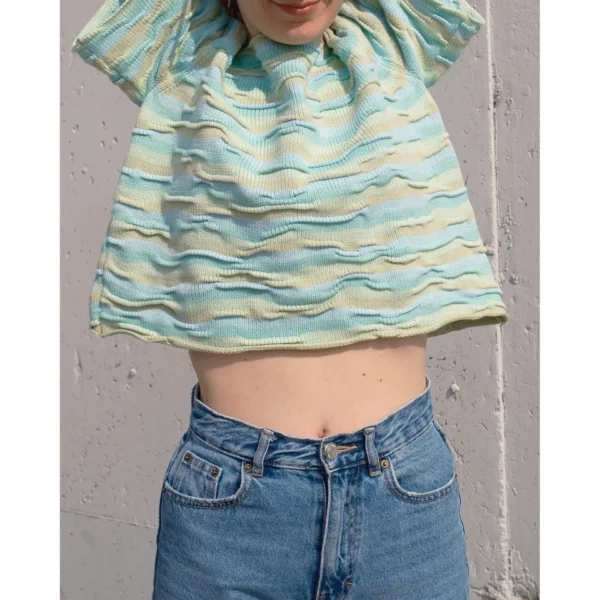 A young woman raises her arms while wearing Breezer (Mint), showcasing the handmade knit t-shirt’s bumpy texture. The unique knitting technique and warm tones of Minty Ice Blue and Muted Olive come to life in this dynamic shot.