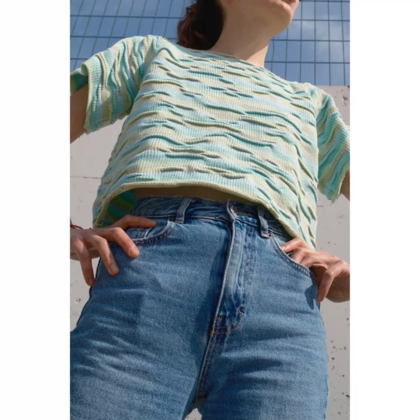 A young woman wears Breezer (Mint) while standing with her hands on her hips, captured from a low angle. The sky frames the shot as the handmade knit t-shirt’s bumpy texture and warm tones of Minty Ice Blue and Muted Olive shine through.