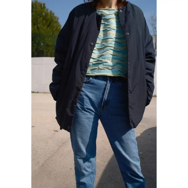 A young woman pairs Breezer (Mint) with a thick, dark jacket, showcasing the handmade knit t-shirt's bumpy texture. The warm tones of Minty Ice Blue and Muted Olive complement the layered look, highlighting the unique craftsmanship of this piece.